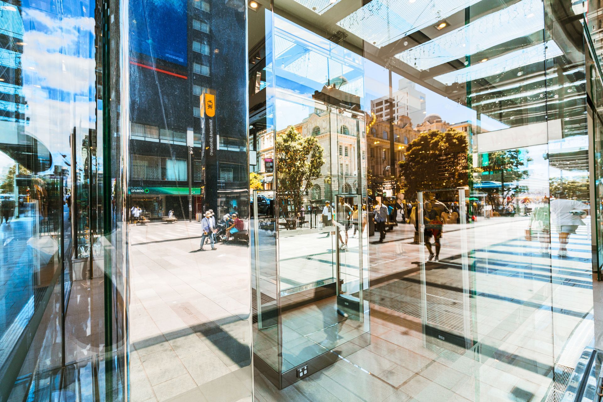 Glass storefront reflects a city street scene: pedestrians, buildings, and trees.