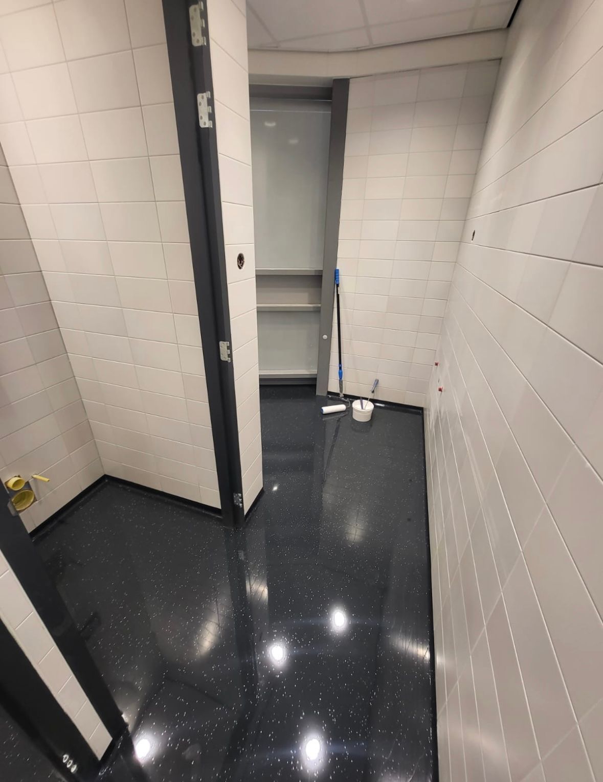 Kleine kamer met een glanzende zwarte vloer en witte tegelwanden; een deuropening leidt naar een smalle bergruimte met planken.