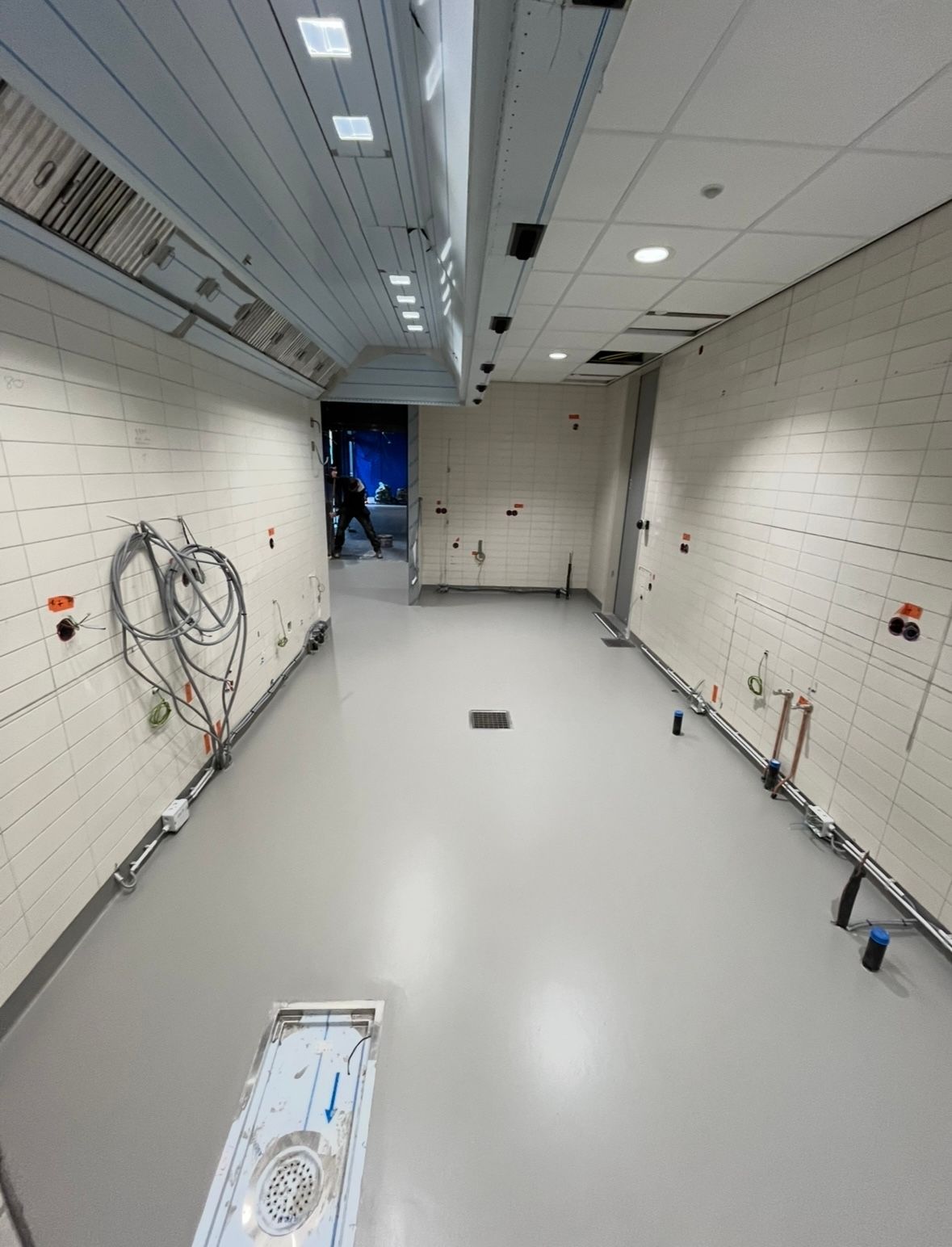 Een lege, langwerpige kamer met witte muren, een grijze vloer en plafondverlichting. Op de achtergrond staat een persoon.