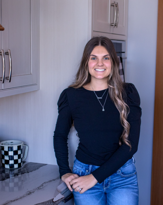 Woman leans against a wall in a kitchen, smiling. She wears a black shirt, jeans, and necklaces.