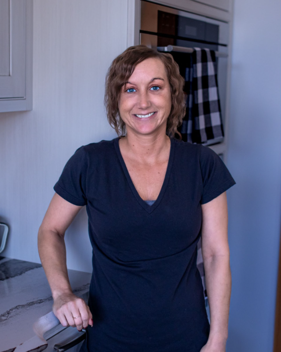 Woman in a dark V-neck shirt smiling in a kitchen with a modern appliance in the background.