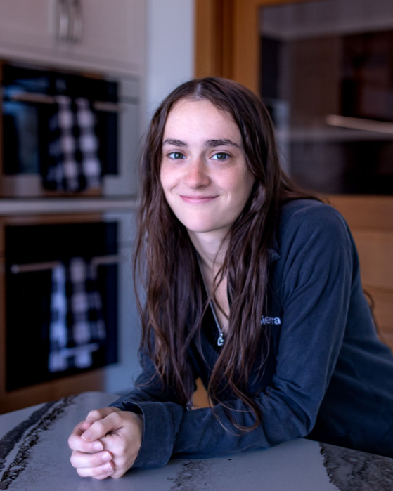 Woman smiling, leaning on a counter, kitchen setting. Long brown hair, dark jacket, hands clasped.