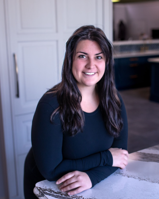 Woman with long dark hair smiles, leaning on a counter, wearing a black top. Kitchen setting with white and blue cabinets.