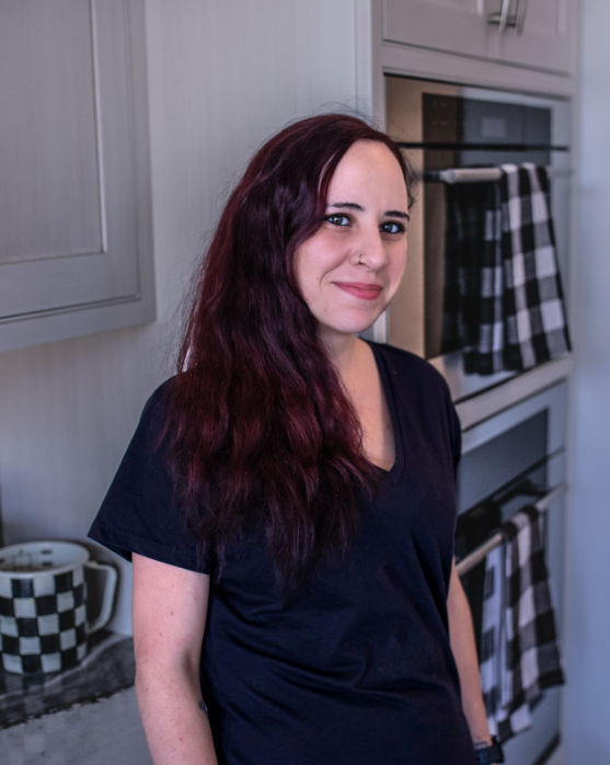 Woman with long red hair, wearing a black shirt, in a kitchen with checkered towel and a mug.