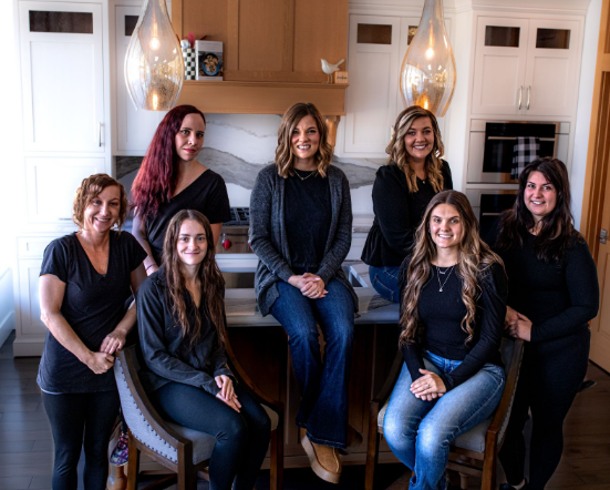 Group of women posing in a kitchen. Some are sitting and standing, all in dark clothing.
