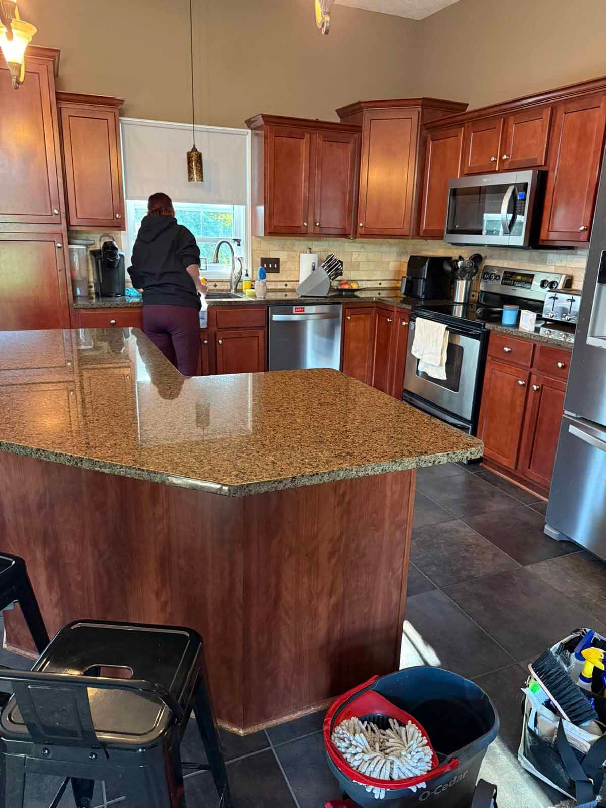 Person in a kitchen with brown cabinets and granite countertops; person near sink washing dishes.