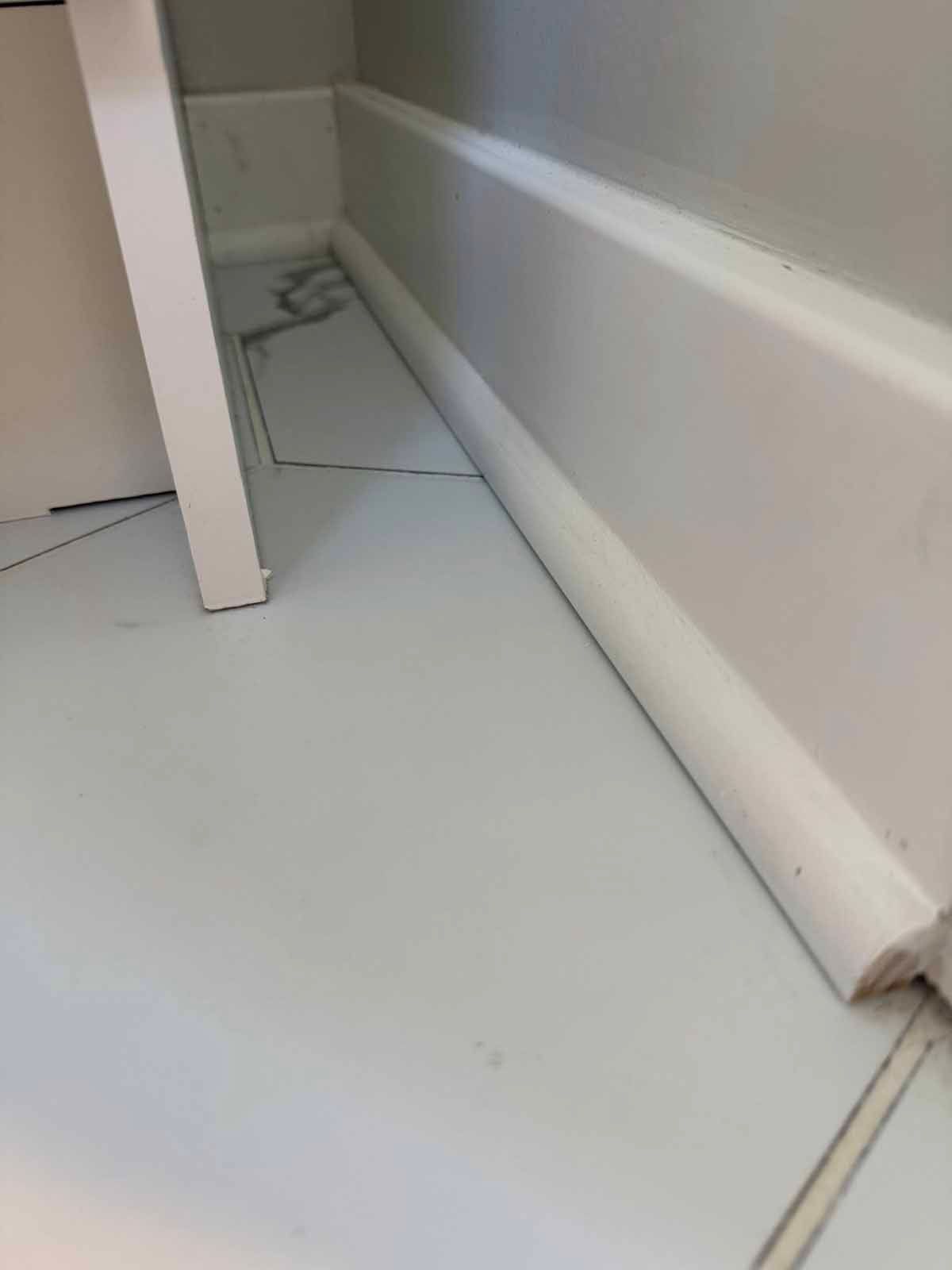 White baseboard against a gray wall, white tile floor, the corner of a white cabinet.