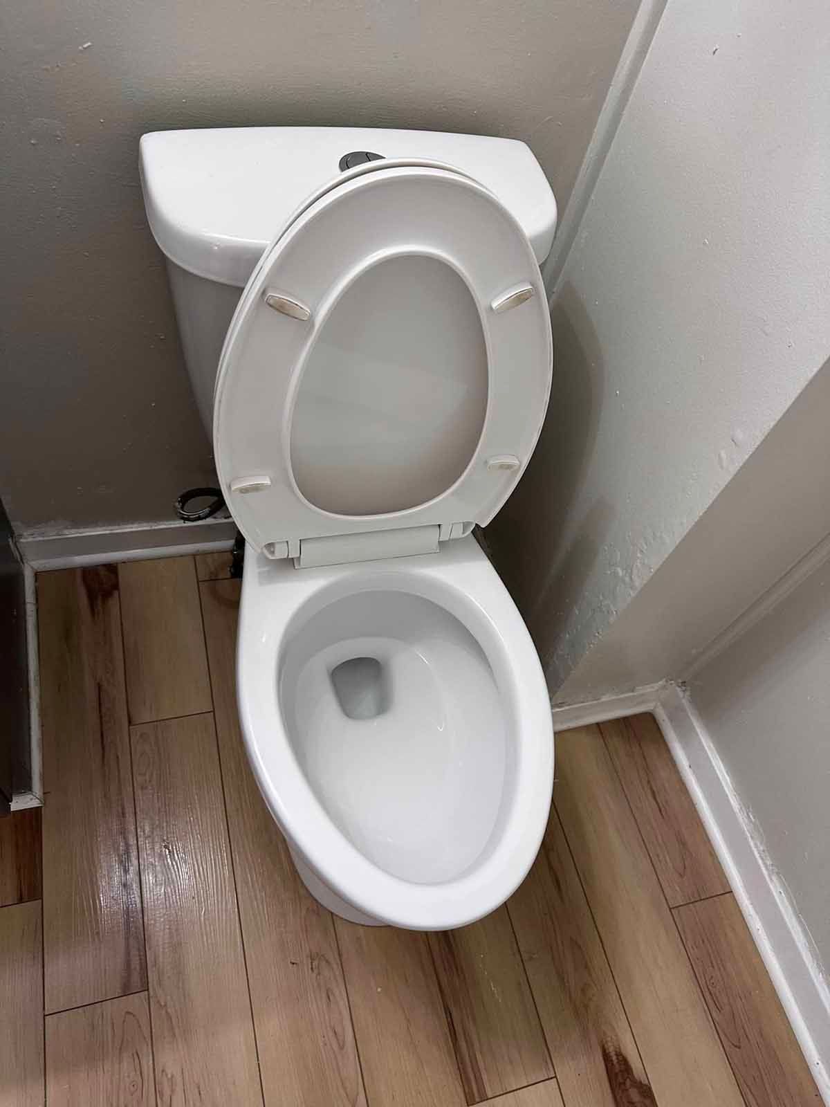 White toilet with seat up, on wood-look floor, in a white-walled room.