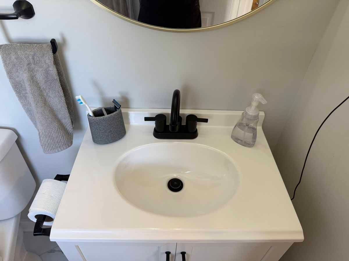 White bathroom sink with black faucet, hand towel, and toiletries. Round mirror overhead.
