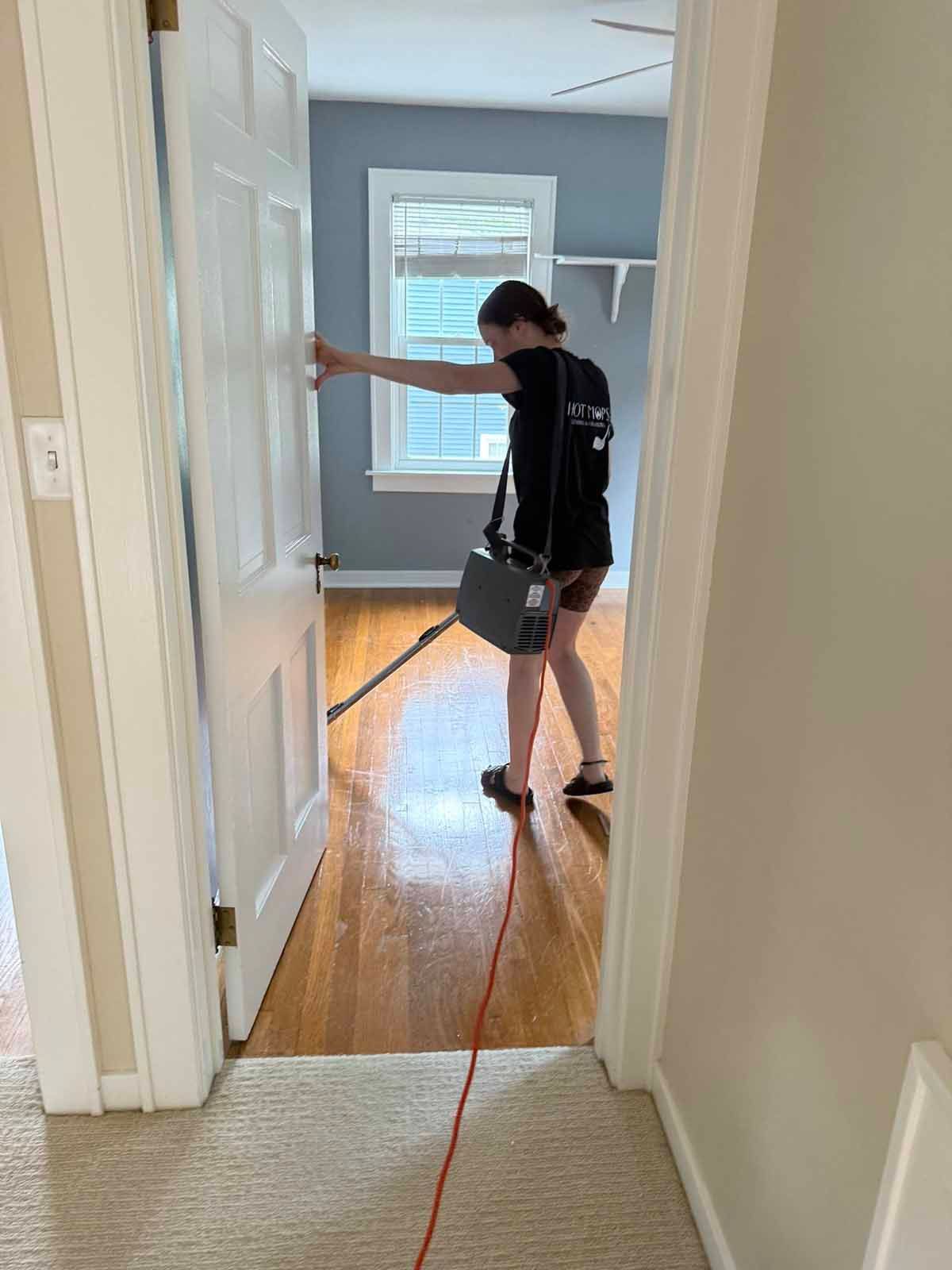 Person vacuuming hardwood floor in a room. Doorway in the foreground. Brightly lit room with blue walls.