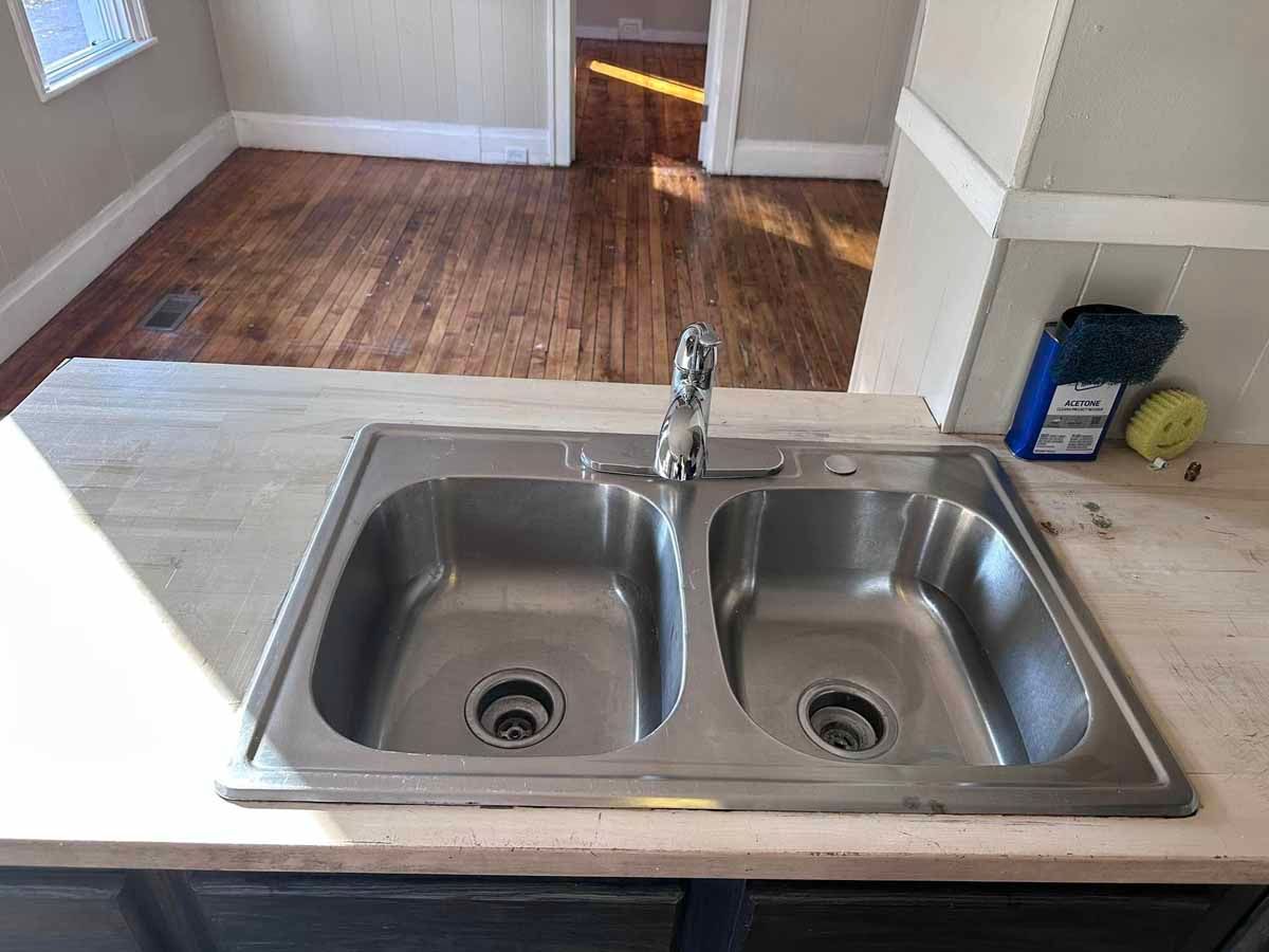 Stainless steel double kitchen sink on a light-colored countertop. Dark wood floors and a doorway are in the background.