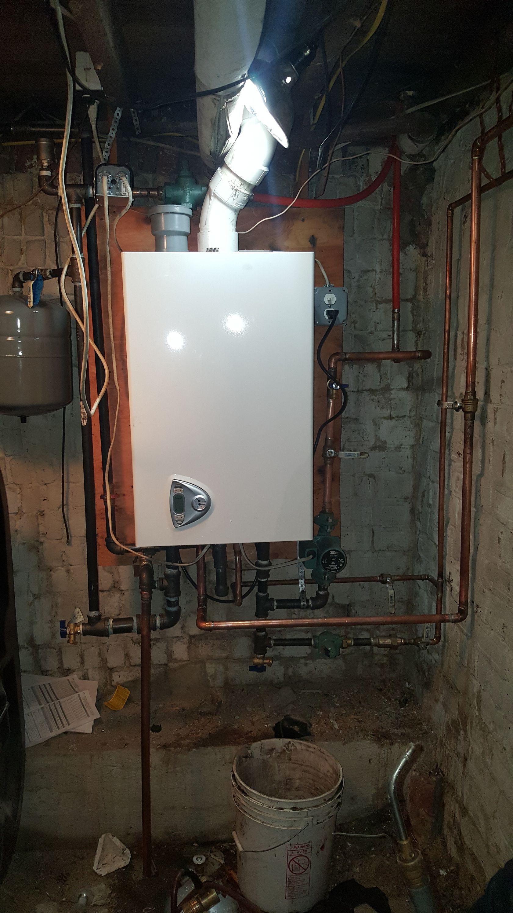 A white boiler is sitting in a basement next to a bucket.