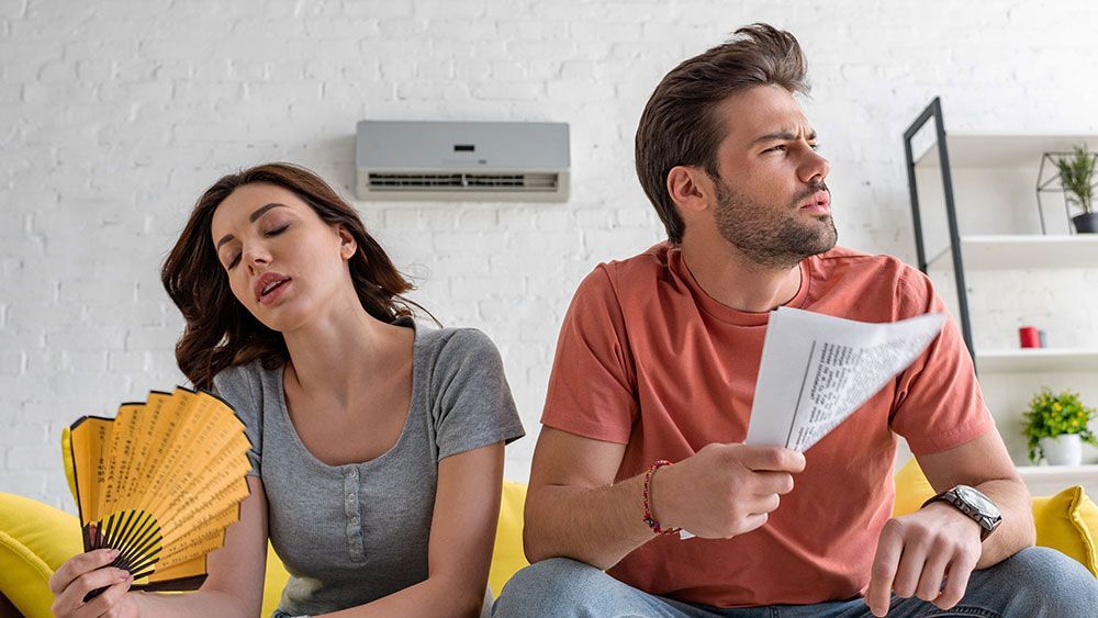 A man and a woman are sitting on a couch under an air conditioner.