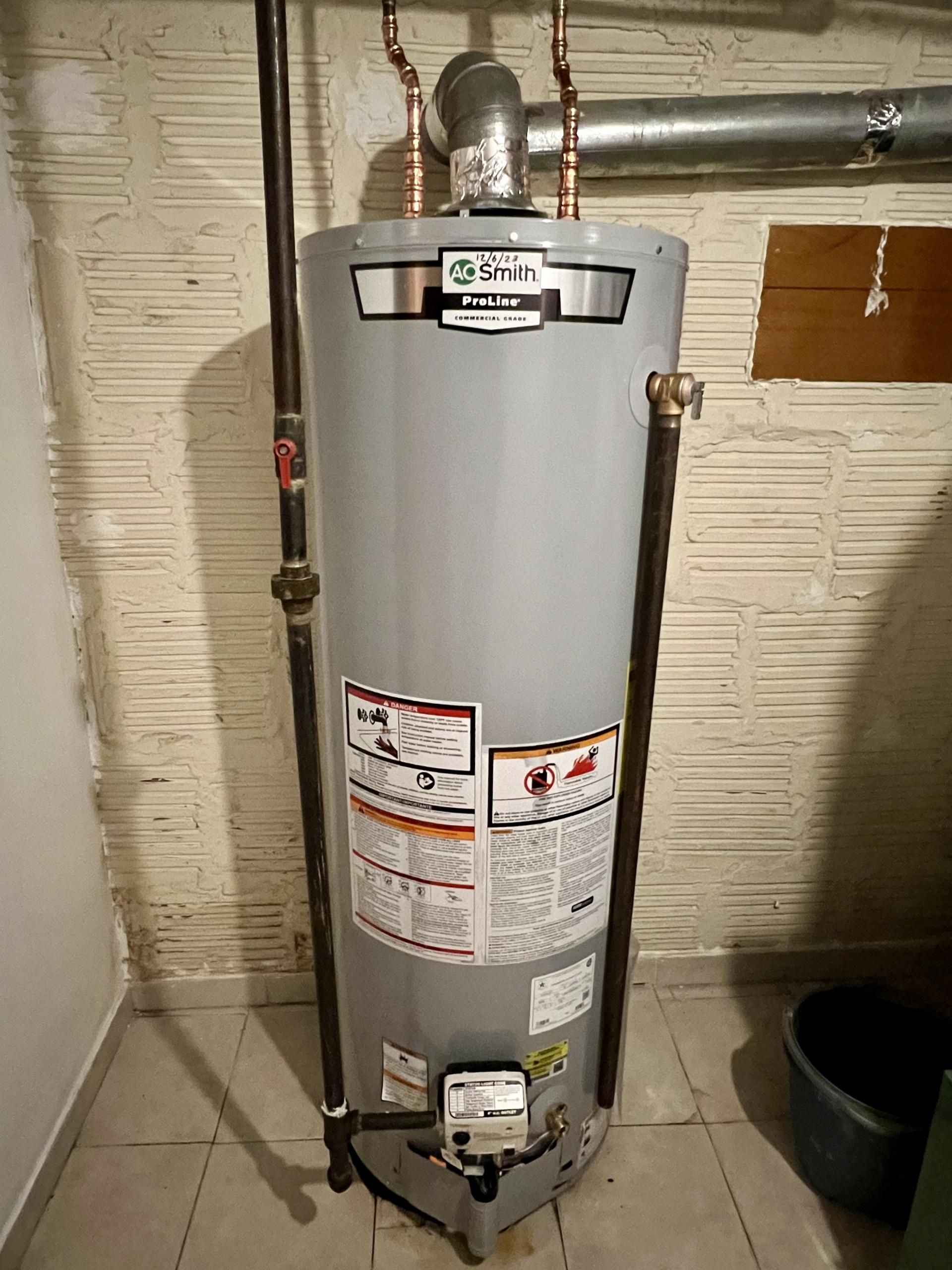 A tall, gray water heater with pipes in a basement corner.