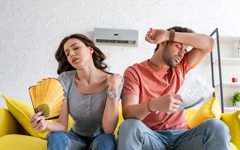 A man and woman are hot and bothered indoors. She fans herself with a yellow fan, he wipes his brow, near an AC unit.
