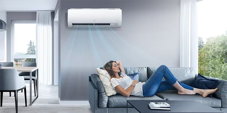 Woman relaxing on a sofa beneath an air conditioner. She is reclining with her eyes closed, with blue air blowing on her.