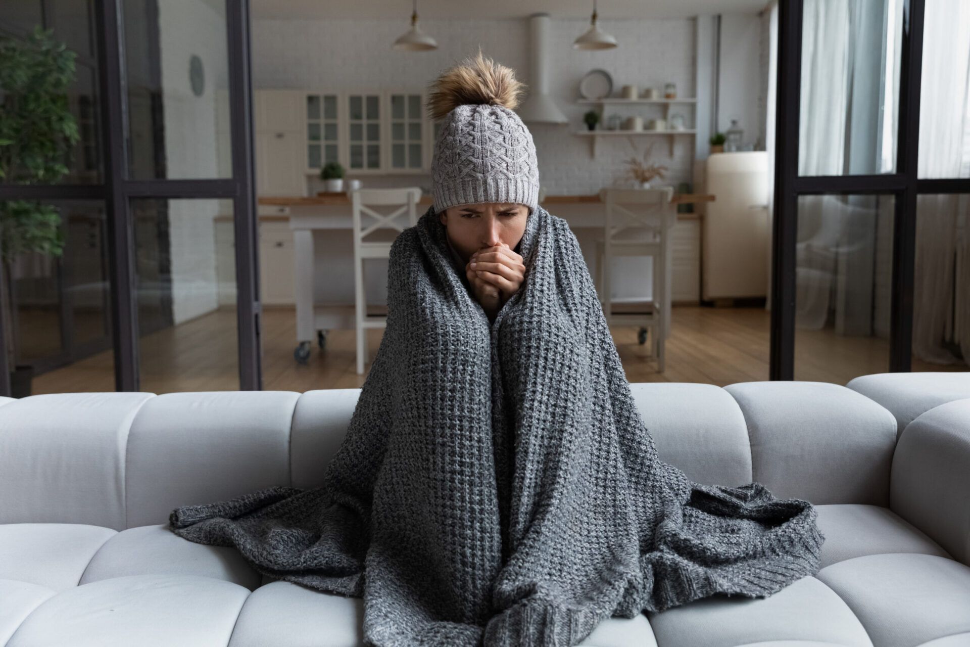 Person wrapped in blanket, wearing hat, shivering on a couch in a cold-looking living room.