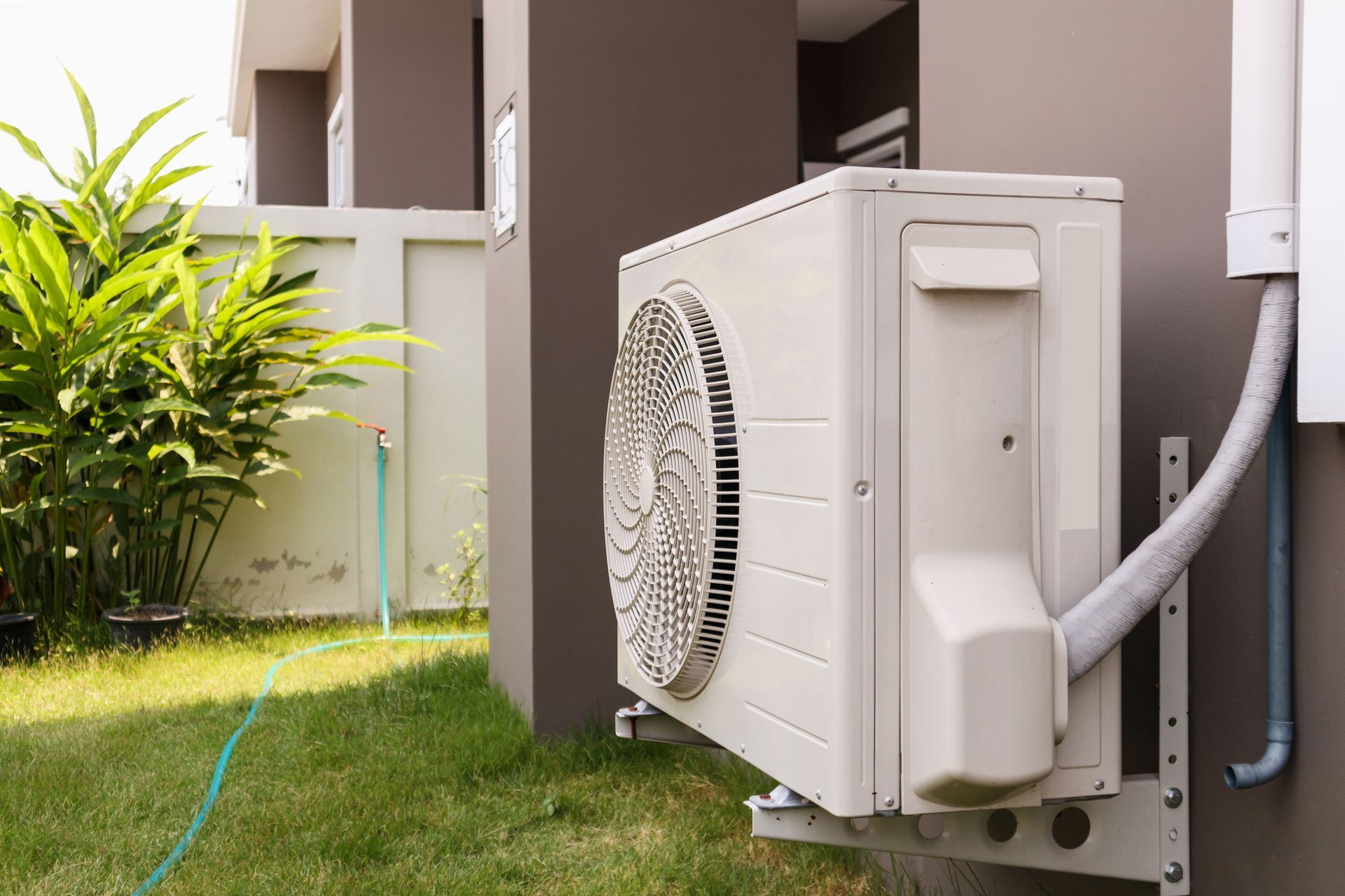 An outdoor air conditioning unit mounted on a wall next to a patch of grass and greenery.