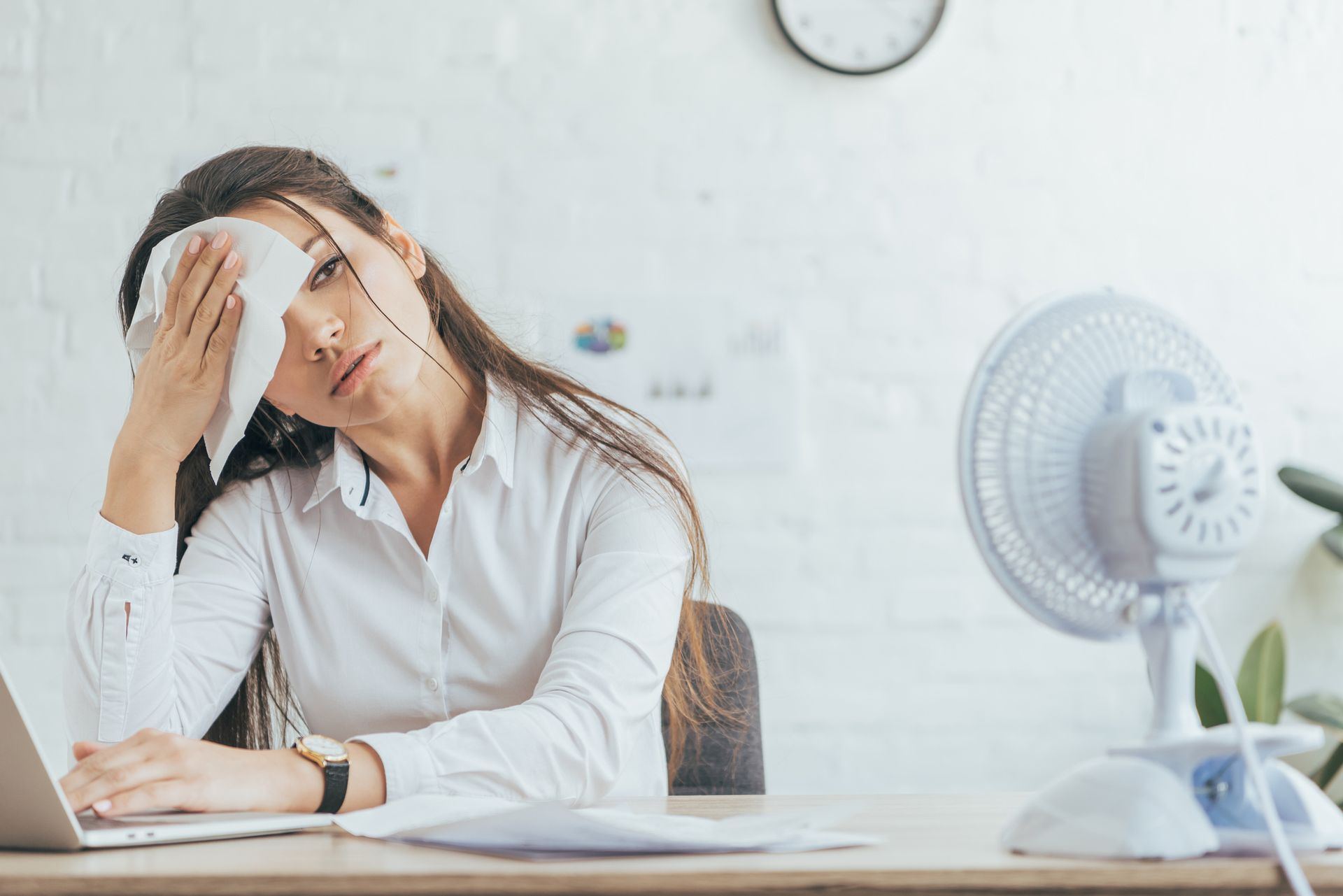 A woman is wiping her face with a towel while sitting at a desk in front of a fan.
