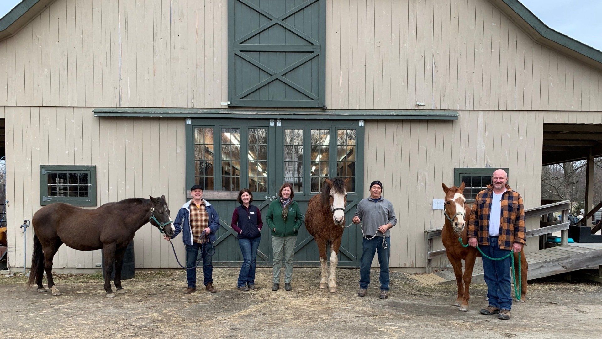 A group of people standing next to horses in front of a barn.