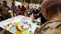 A group of people are standing around a table while a man paints on it.