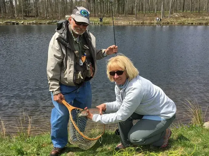 Two people kneel by a pond, holding a fish in a net near the water’s edge.