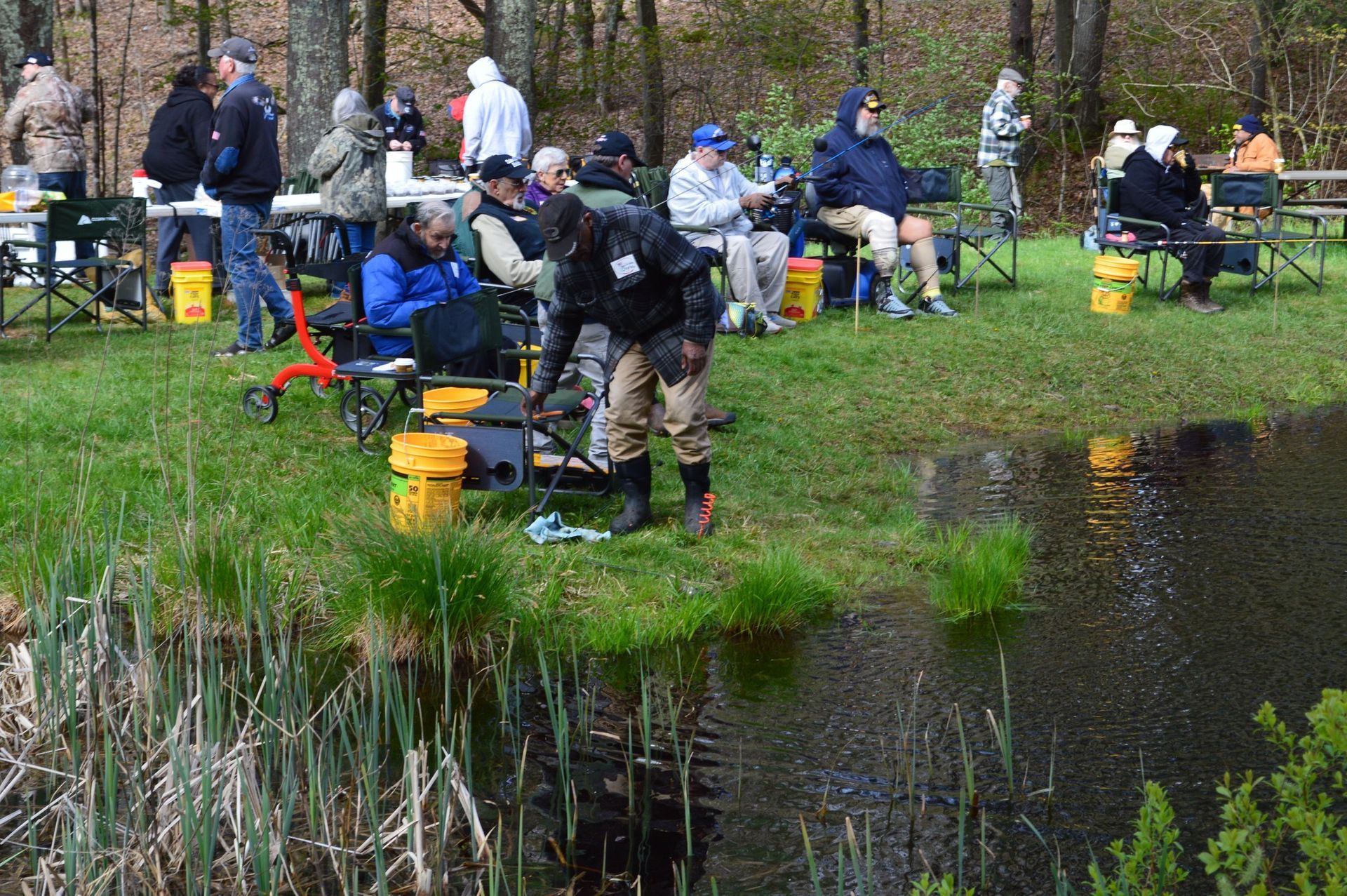 People fishing along a grassy pond bank with gear and buckets on a cloudy day