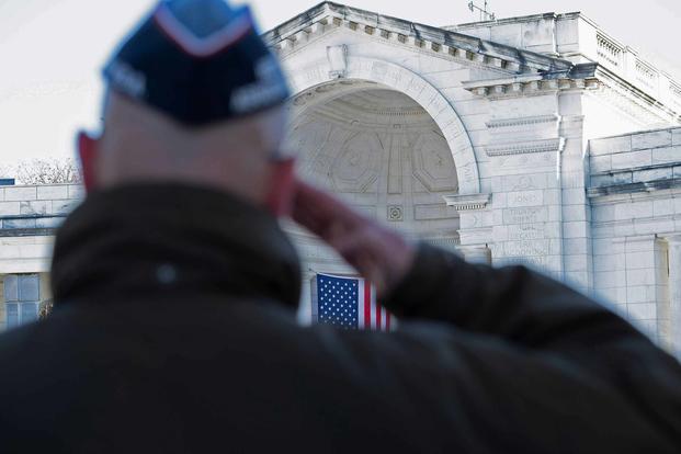 A man is saluting in front of a building with an american flag in the background.