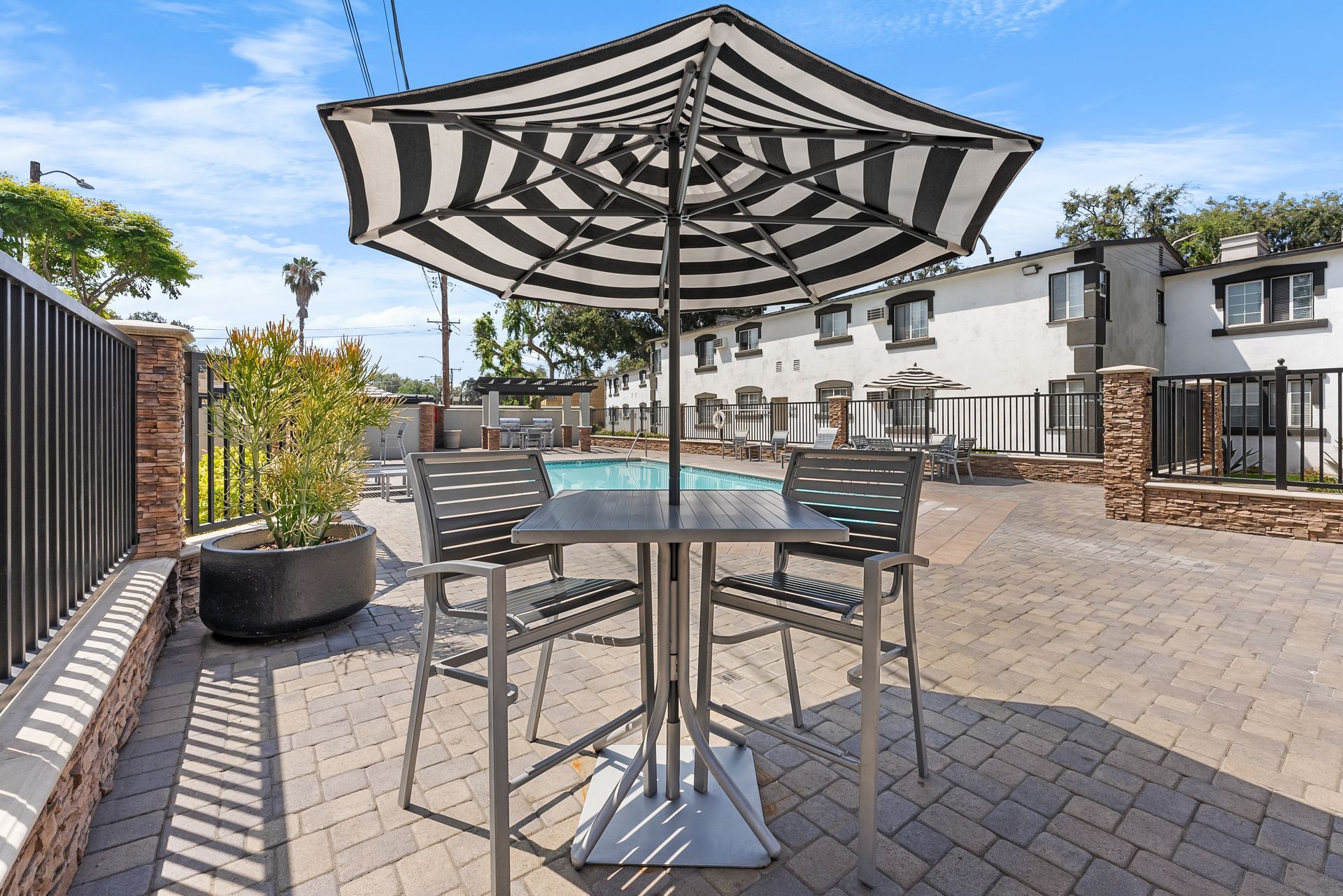 Patio area with pool. Black and white umbrella, table, chairs. Apartments in background.