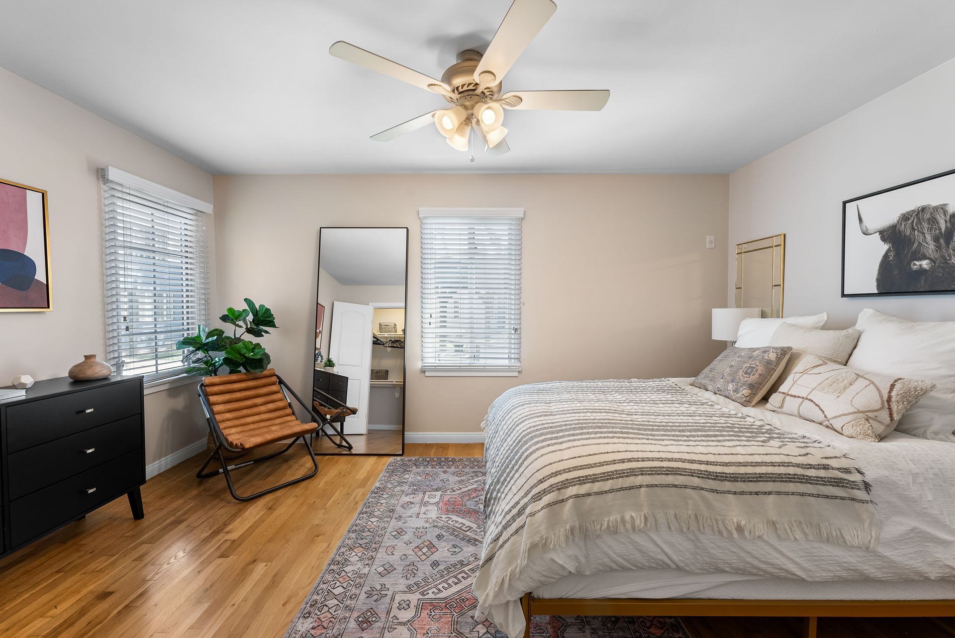 Bedroom with wooden floor, bed with decorative pillows, black dresser, full-length mirror, chair, rug, and art.