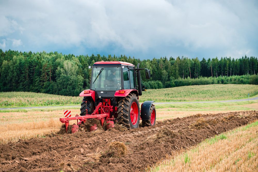 A Red Tractor is Plowing a Field on a Cloudy Day — SMH Valley Fencing & Maintenance In Rutherford, NSW