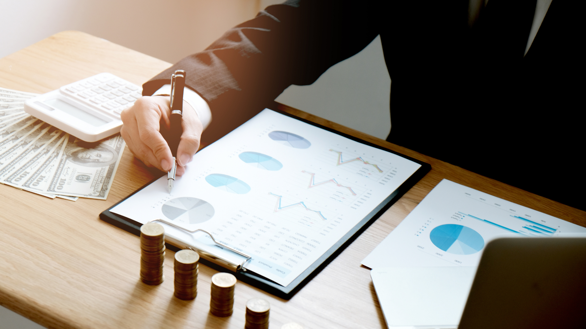 Person in suit analyzes financial charts, with money, coins, and a calculator on a desk.