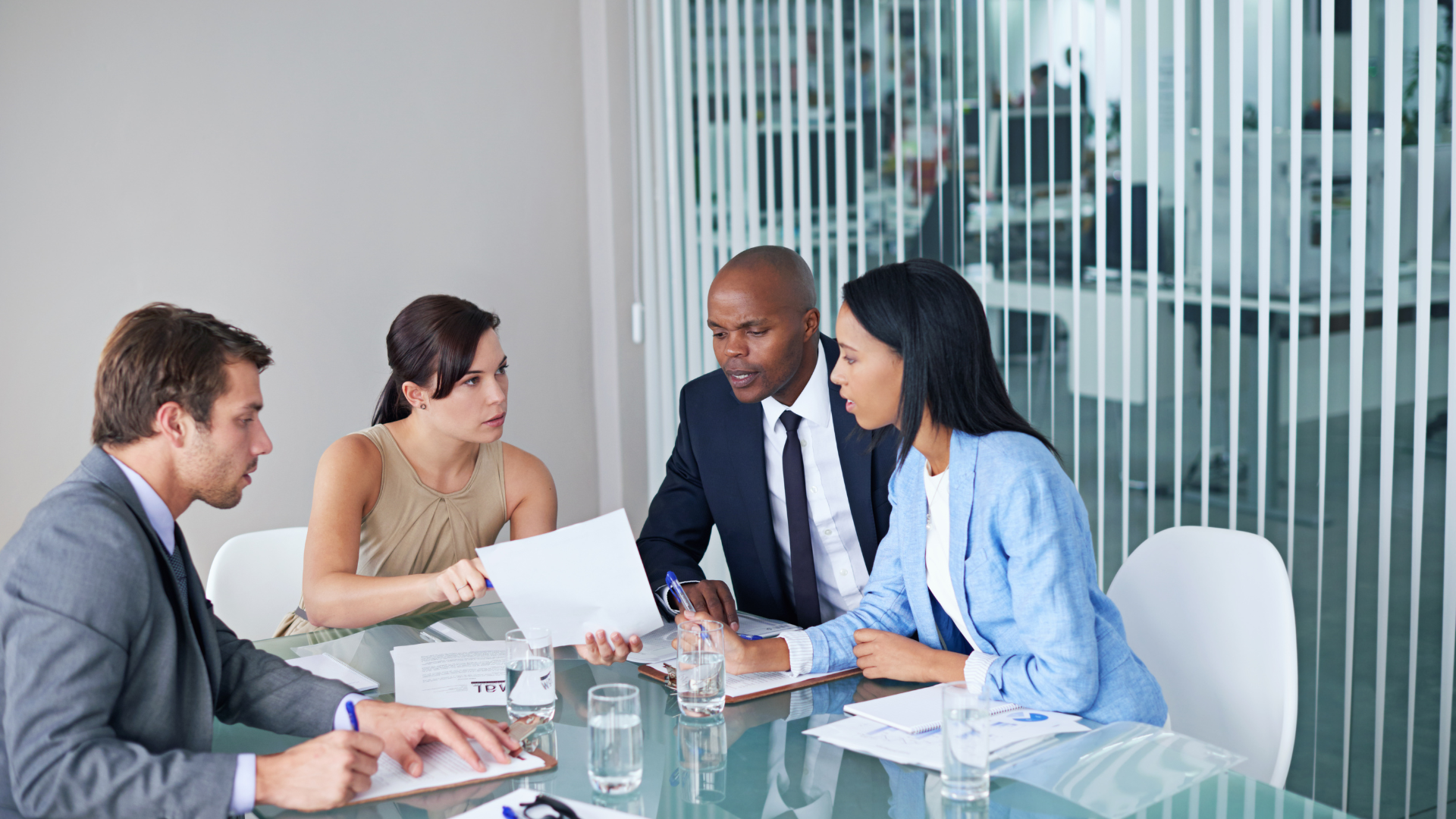 Four businesspeople in a modern office reviewing documents at a glass table.