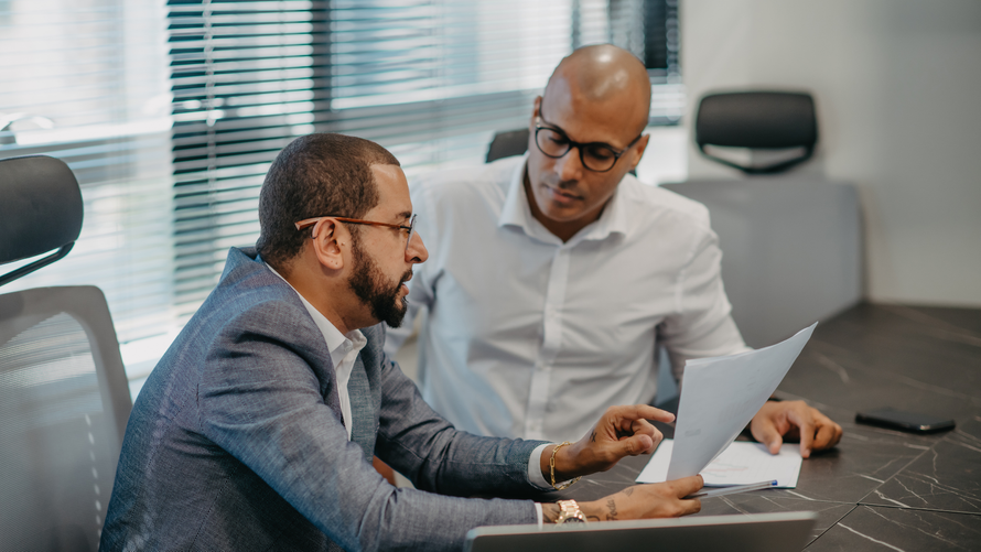 Two men in suits reviewing papers at a table in a modern office.