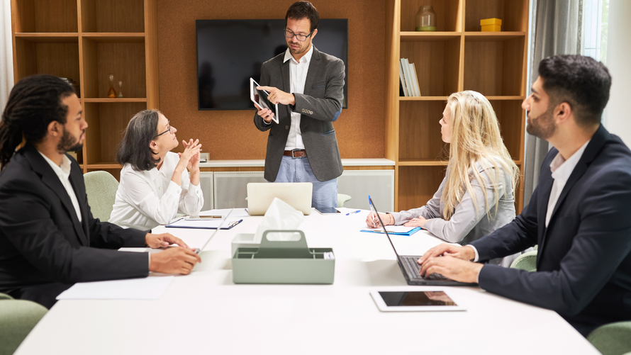 A business meeting in a modern office. A man presents while others listen, taking notes and working on laptops.