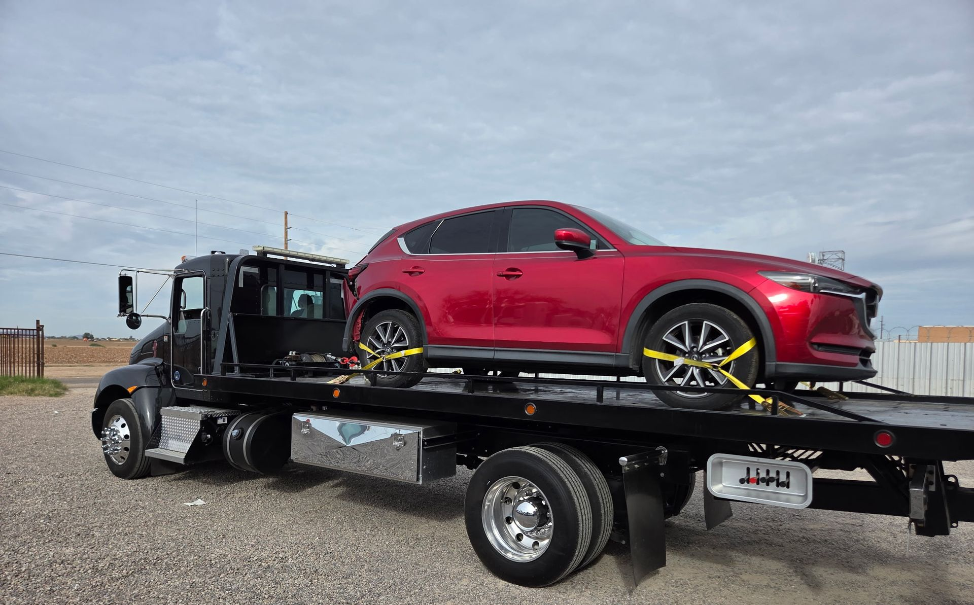 Red SUV secured on a black flatbed tow truck. The truck is parked outdoors on gravel.