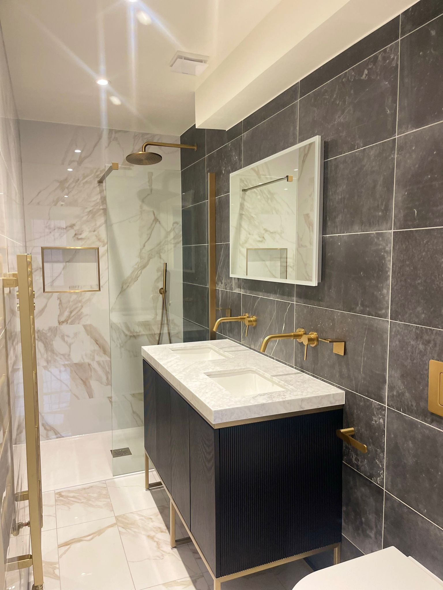 Modern bathroom with marble shower, dark tile walls, and a white vanity sink with gold fixtures