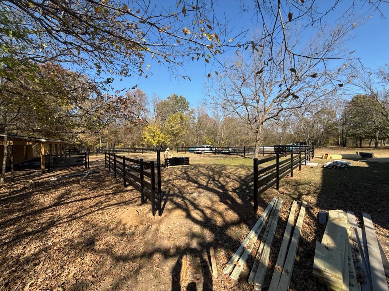 Black wooden fence being built on a brown, leaf-covered lawn in a wooded area on a sunny day.