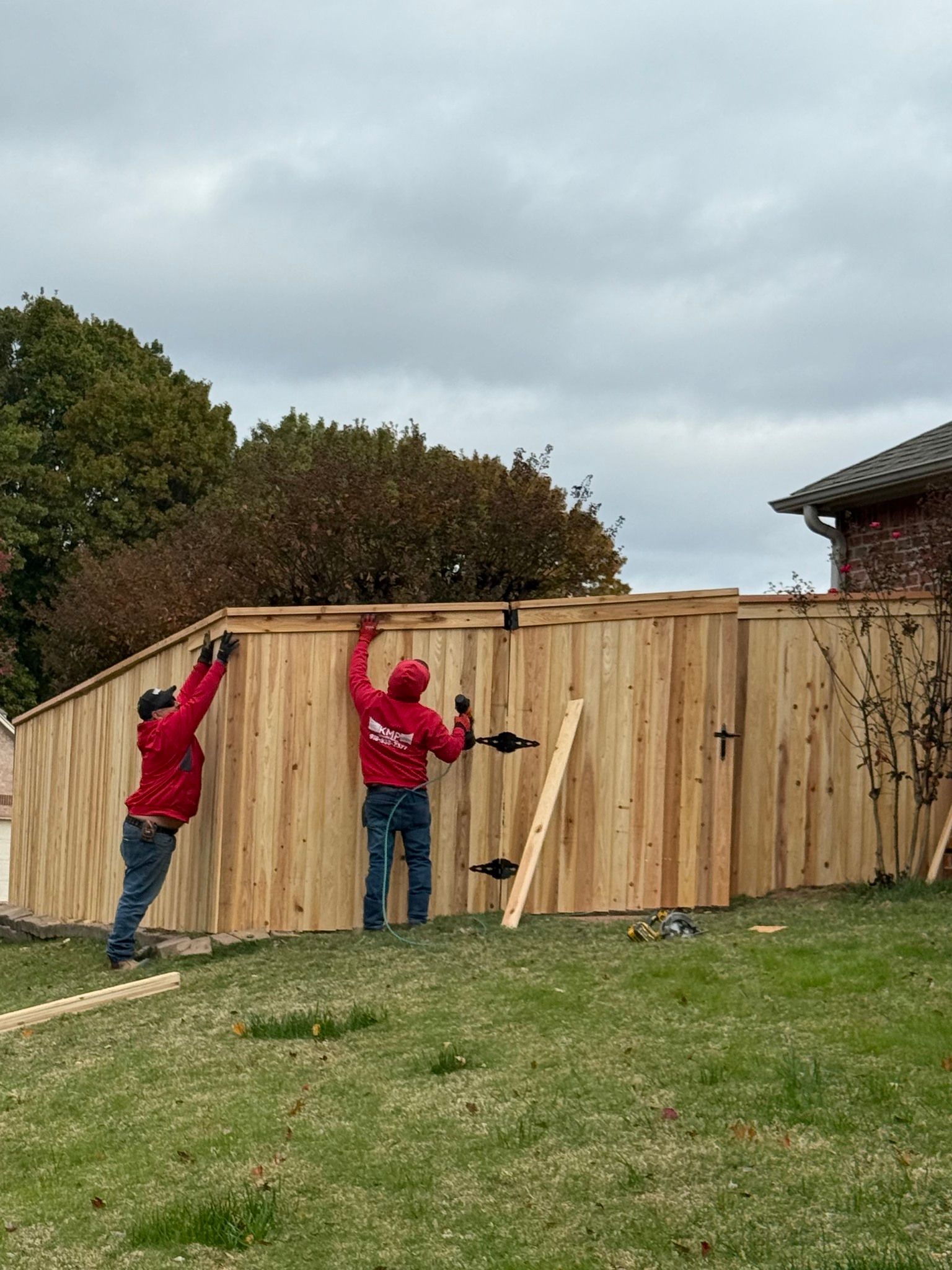Two people in red shirts installing a wooden fence outdoors on a cloudy day.