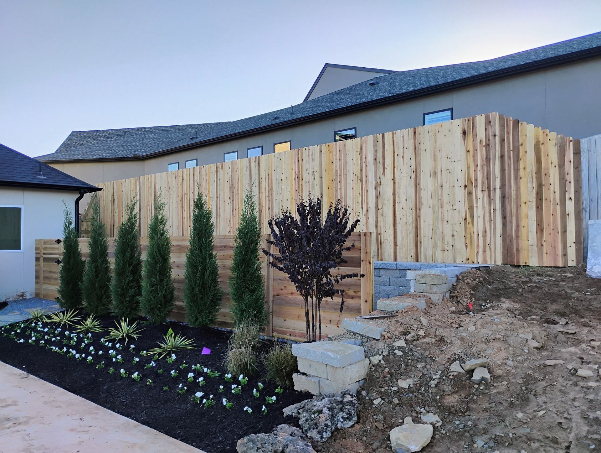 Wooden fence in backyard, with landscaping and small trees. House in background.