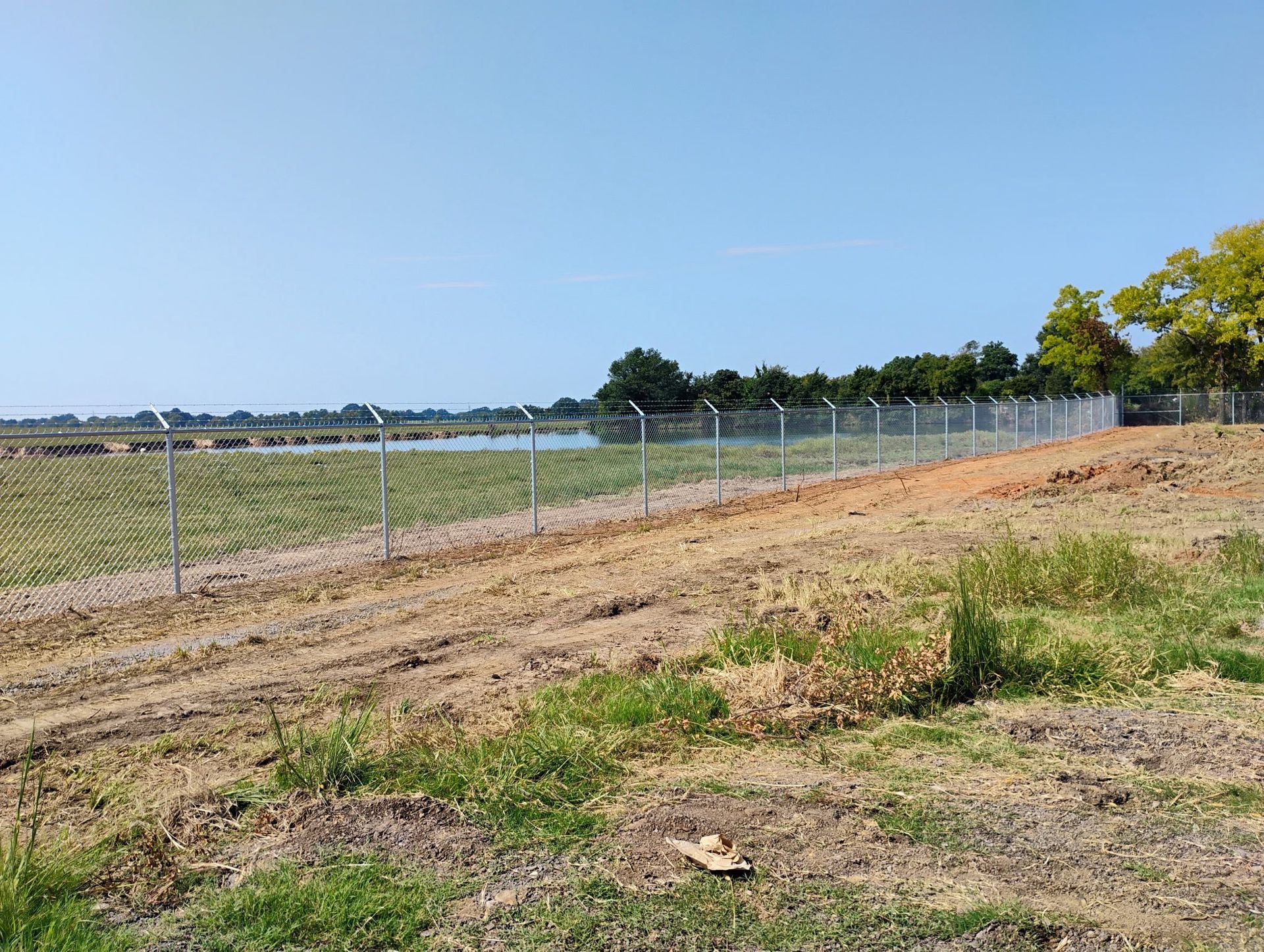 Chain-link fence with barbed wire atop, separating a field from a body of water under a clear sky.