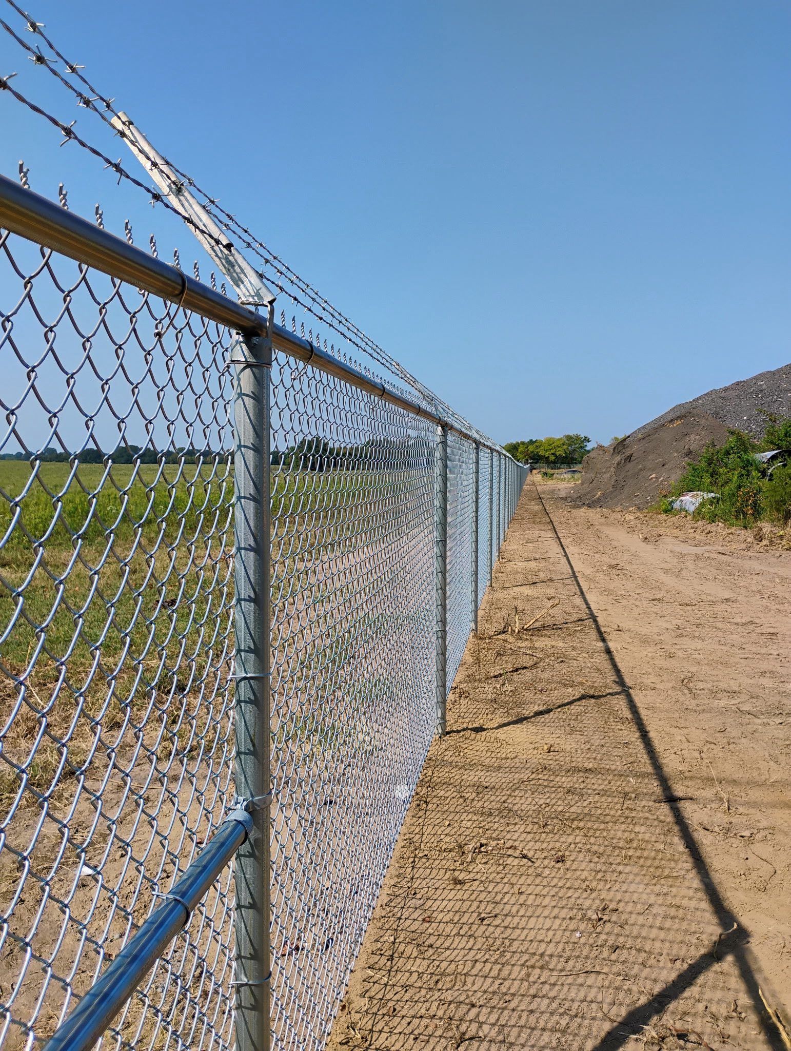 Chain-link fence with barbed wire atop, bordering a dirt path and field under a blue sky.