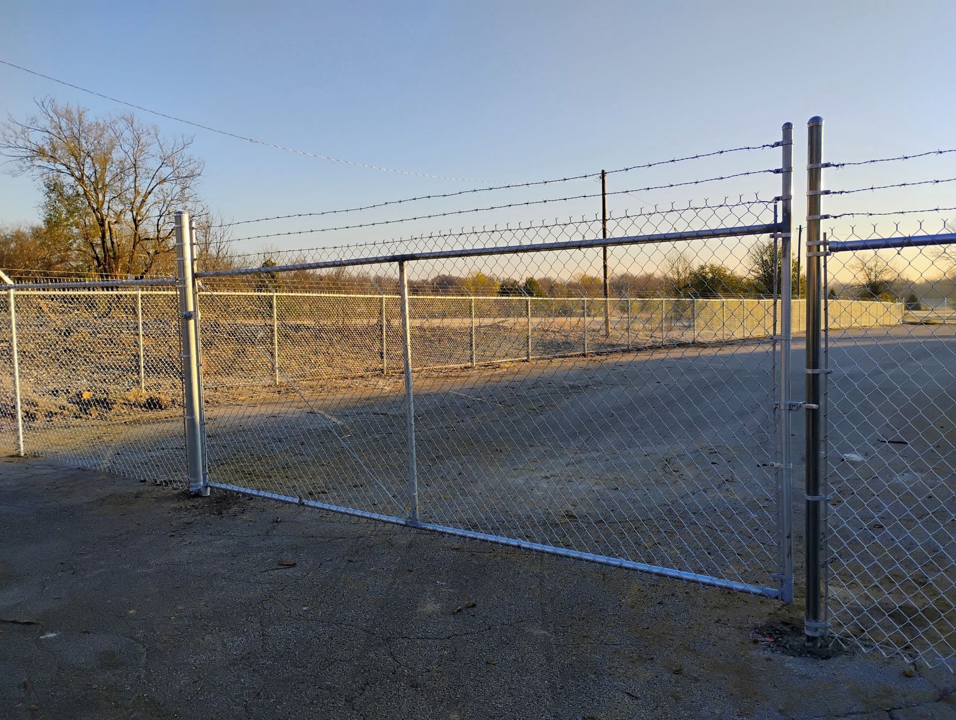 Chain-link fence with barbed wire on top, enclosing a gravel area and field, under a clear sky.