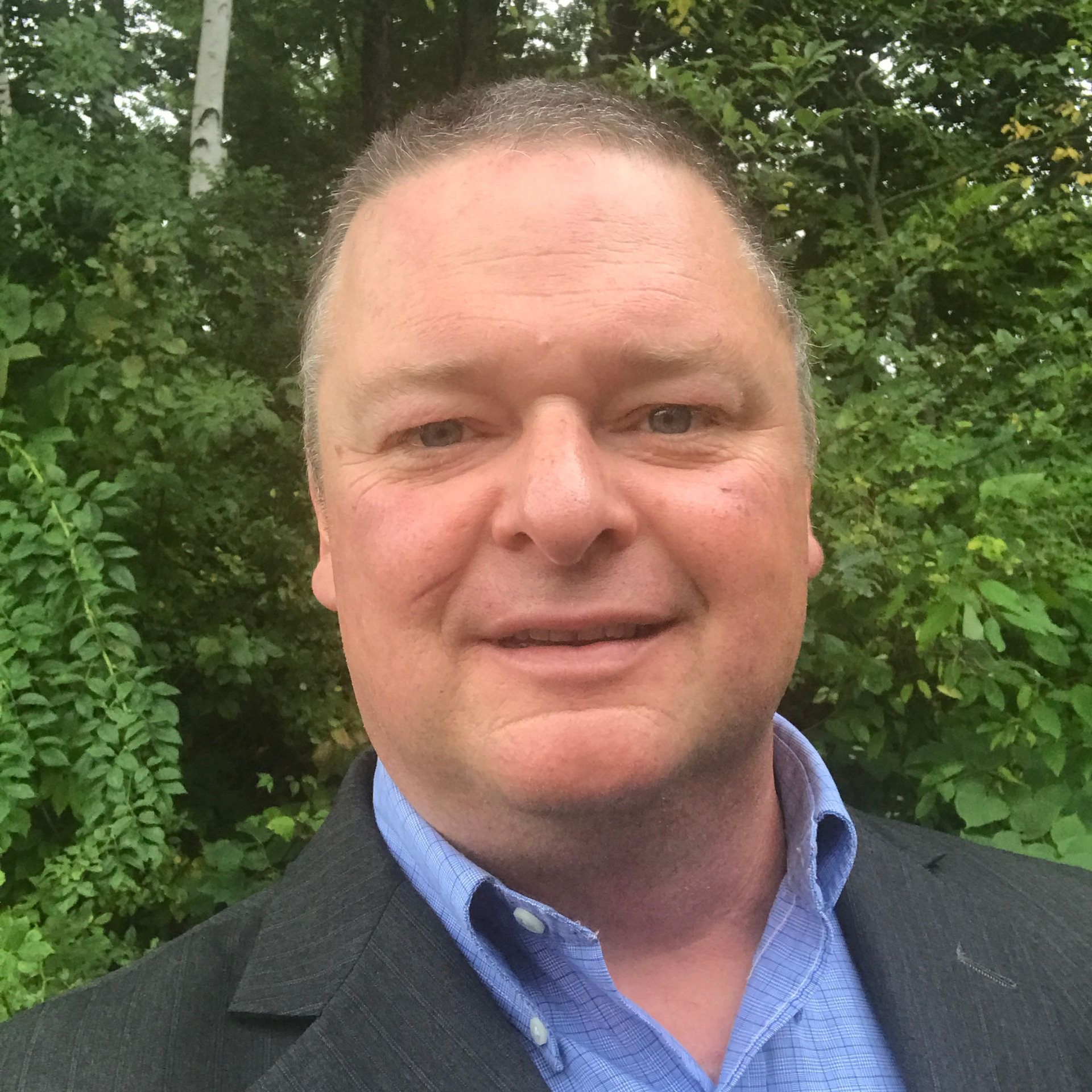 A smiling headshot for the bio of kevin bernard with a background of  trees and bushes.