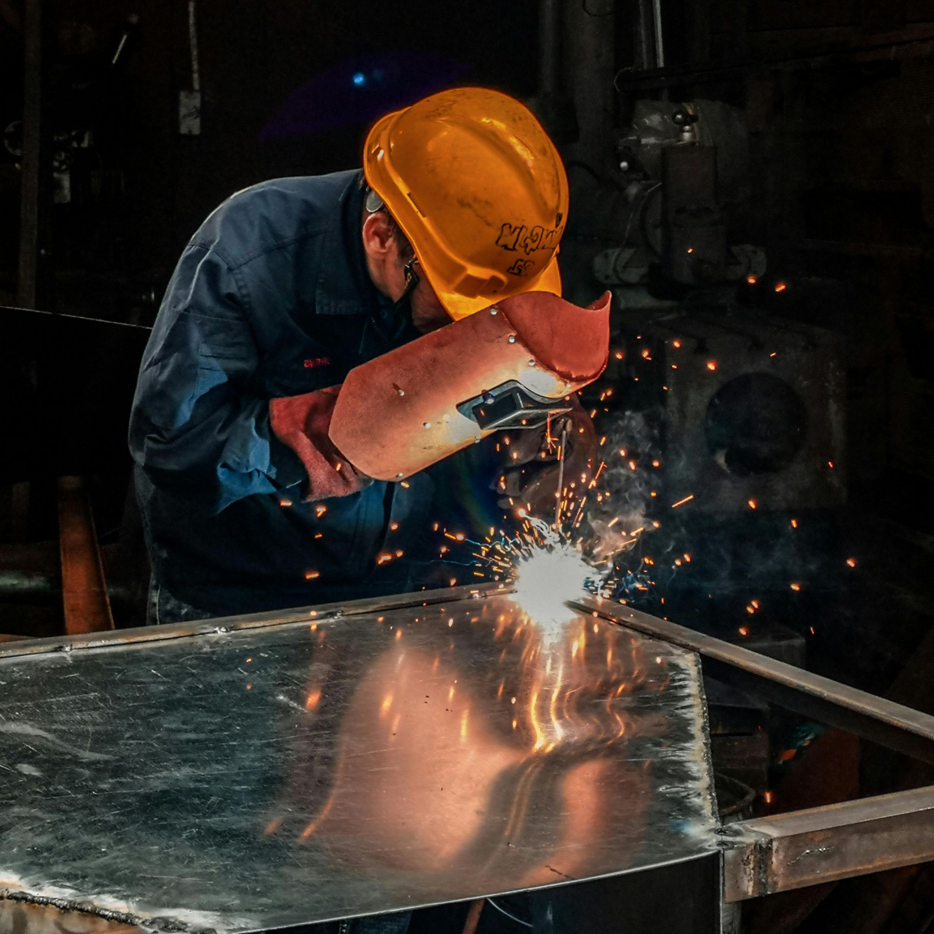 A man welding a piece of metal with sparks wearing a hard hat and welding helmet.