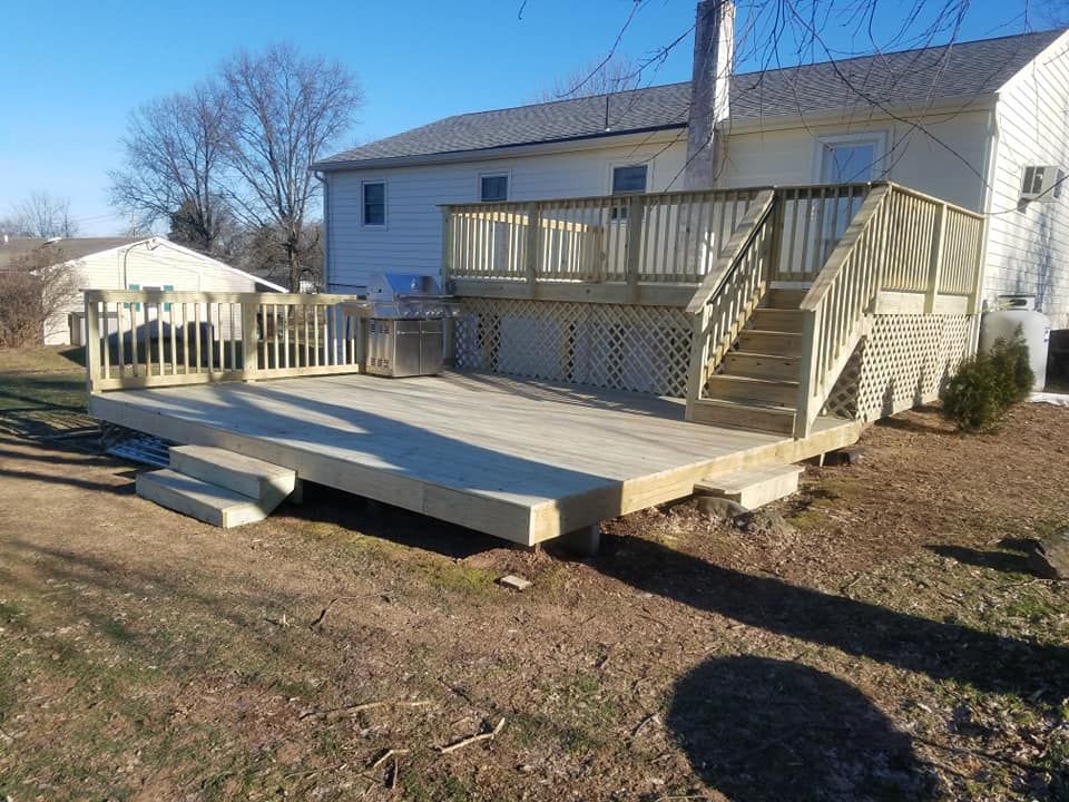 Large wooden deck with stairs attached to a two-story house, set in a grassy yard under a blue sky.