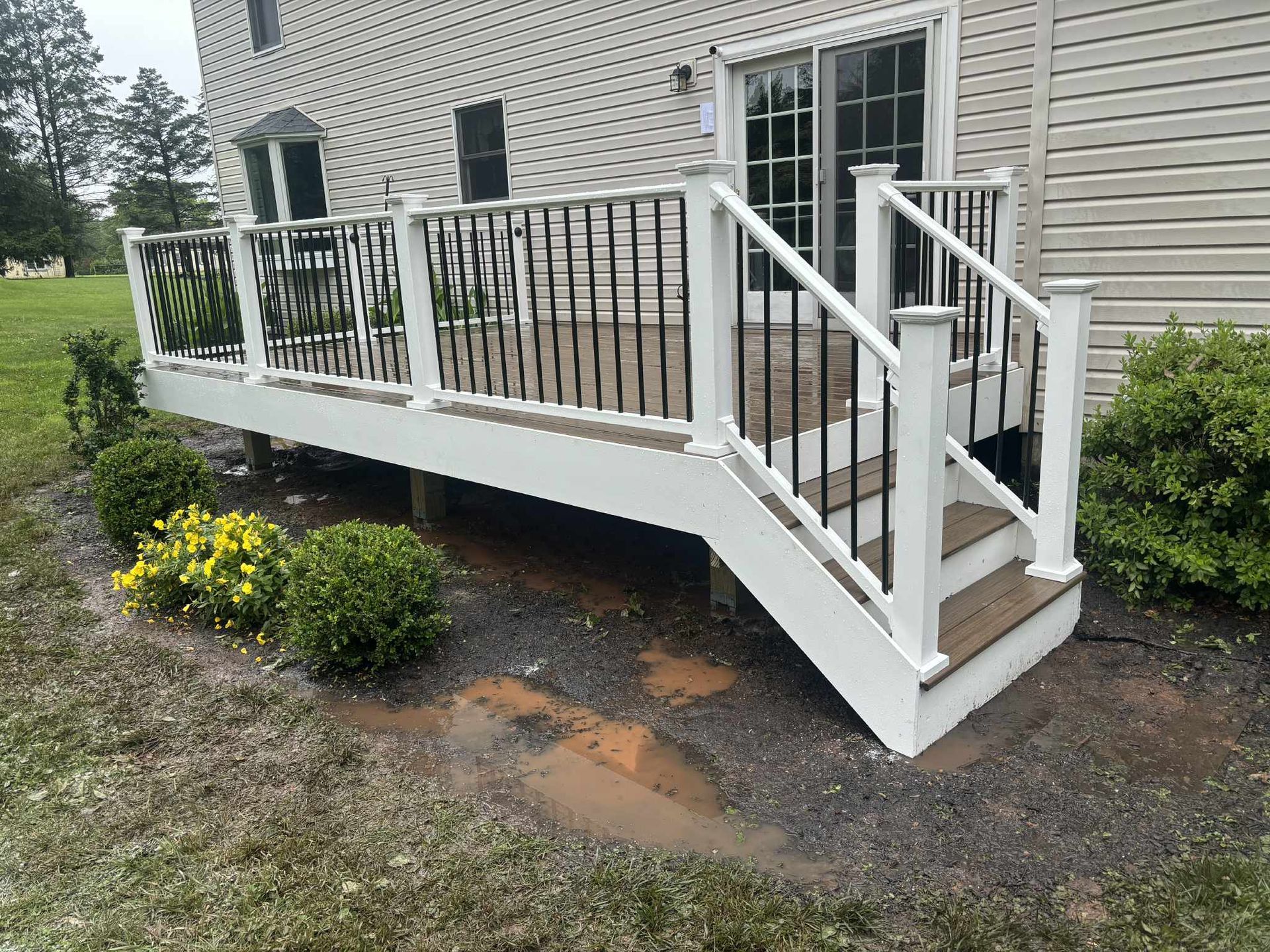 White deck with black railings and steps leading to a sliding glass door, surrounded by landscaping and lawn.