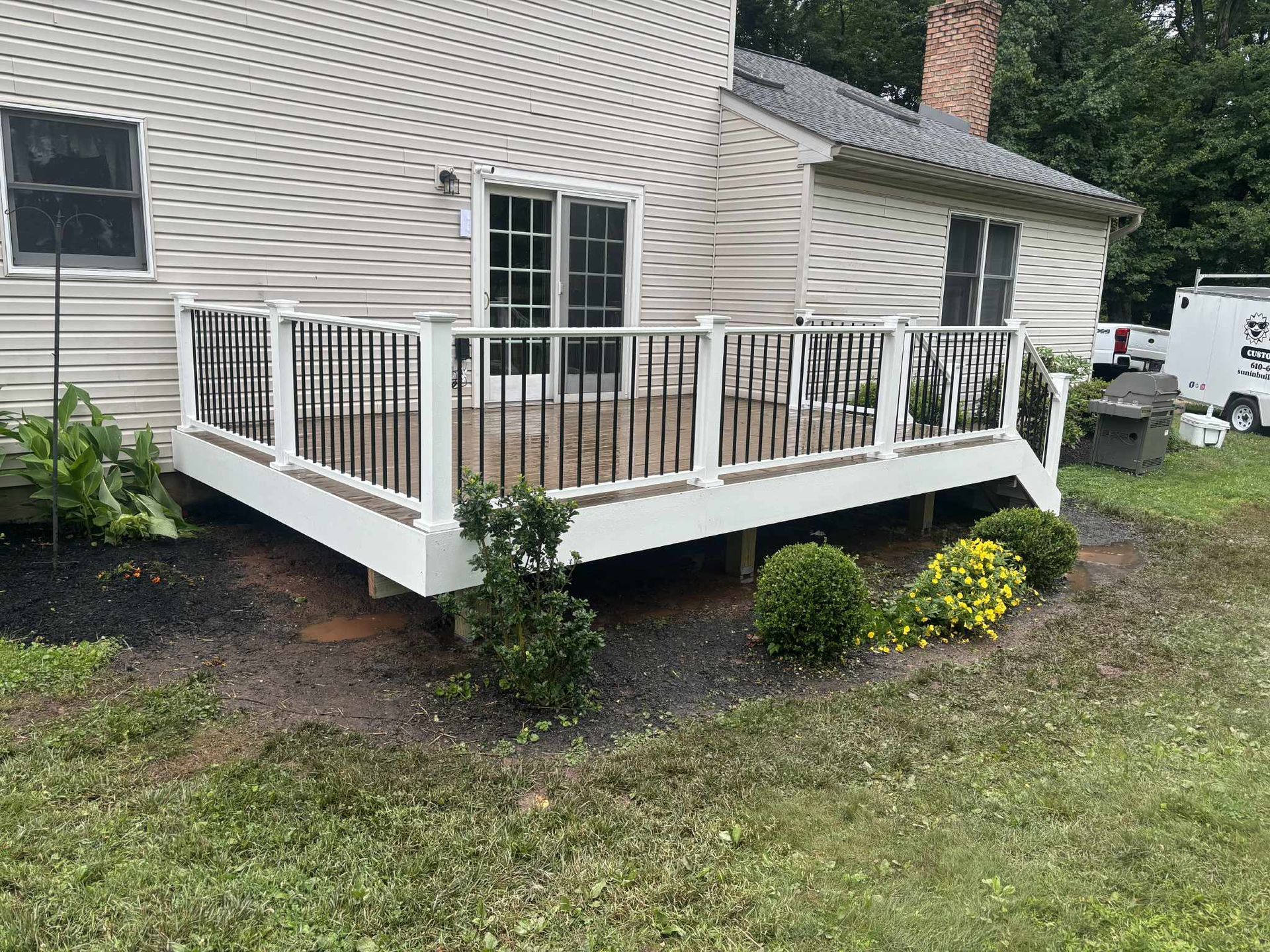 White deck with black railing attached to a beige house, with landscaping and a grill.