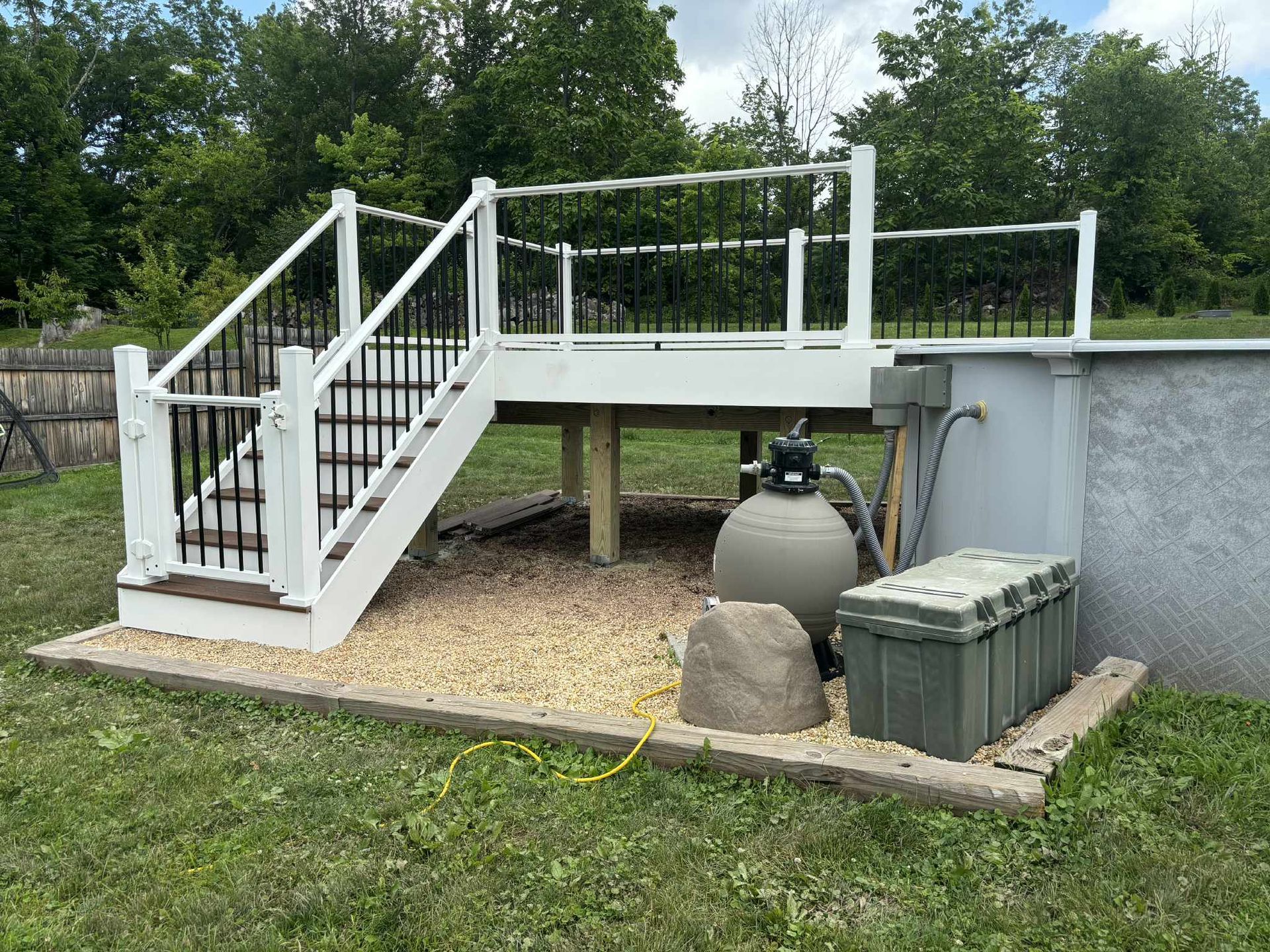 Deck with white railings, black balusters, and stairs, over a gravel base beside an above-ground pool.