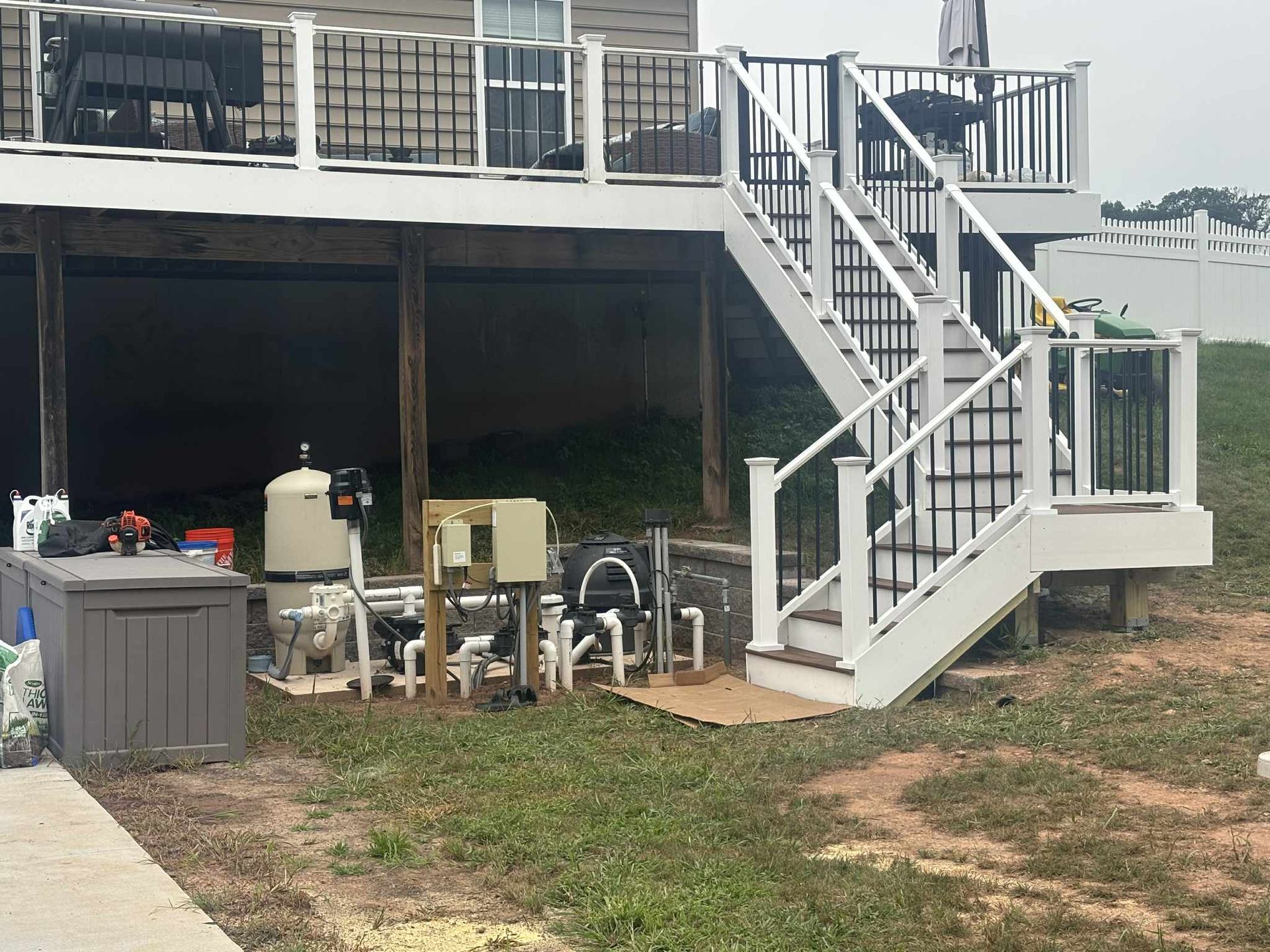 Pool equipment set under a raised deck, with stairs leading up, on a grassy yard.